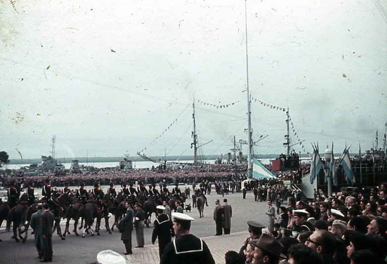 Desfile de caballería junto al río con banderas argentinas y buques engalanados.