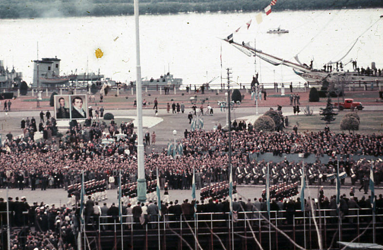 Militares de gala desfilando ante las graderías colmadas de público, con buques ARA La Argentina y ARA Presidente Sarmiento sobre el Río Paraná, Rosario, 1957.