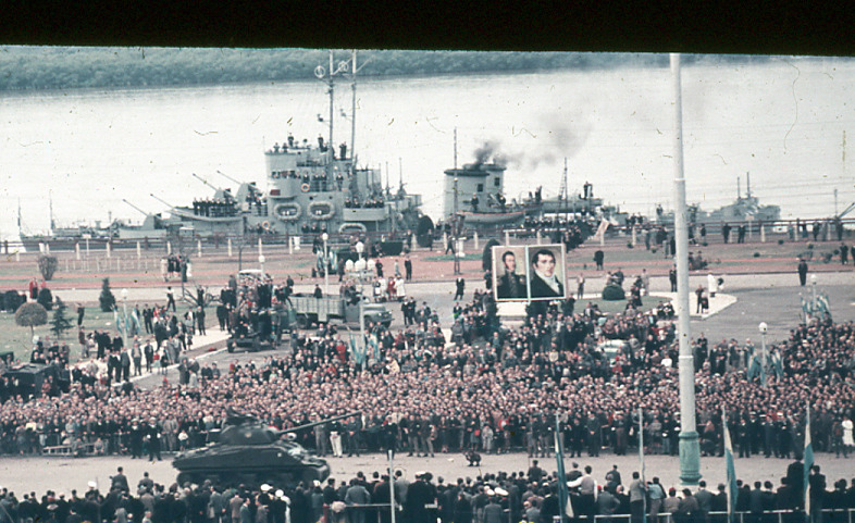 Vehículo militar blindado desfilando ante la multitud, con retratos de los próceres y el buques de la Armada ARA La Argentina sobre el Río Paraná, Rosario, 1957.